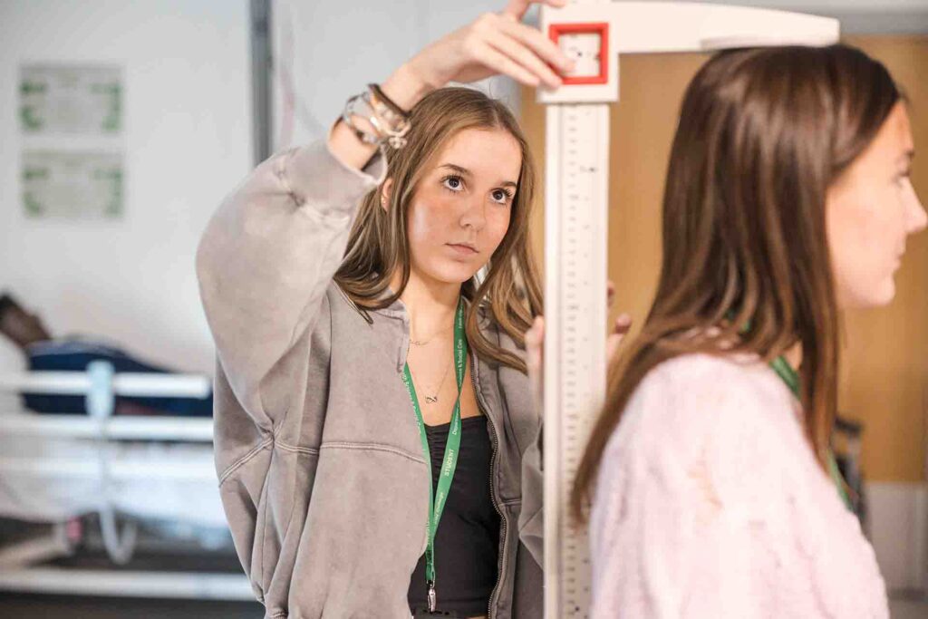 A female student in a grey jacket adjusts a stadiometer height measuring rod over another student's head during a clinical skills exercise
