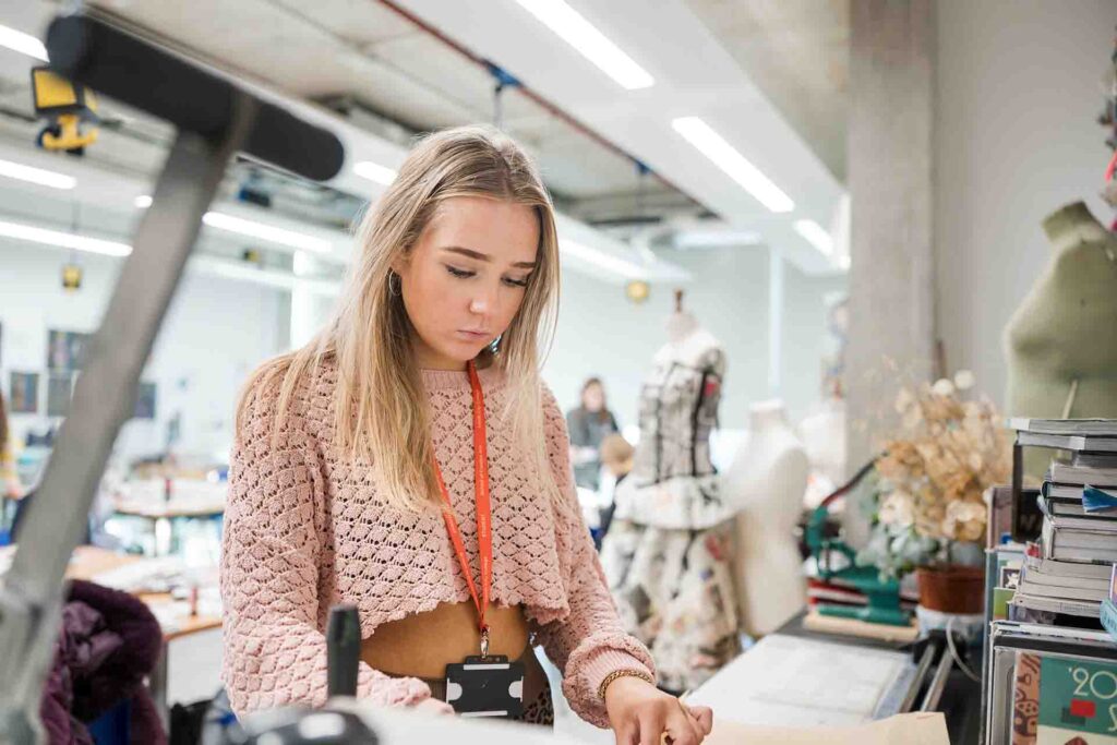 A young blonde female student wearing a pink open-knit cropped sweater and an orange student lanyard focused on her work at a table in a fashion design studio, with mannequins and design books visible in the background