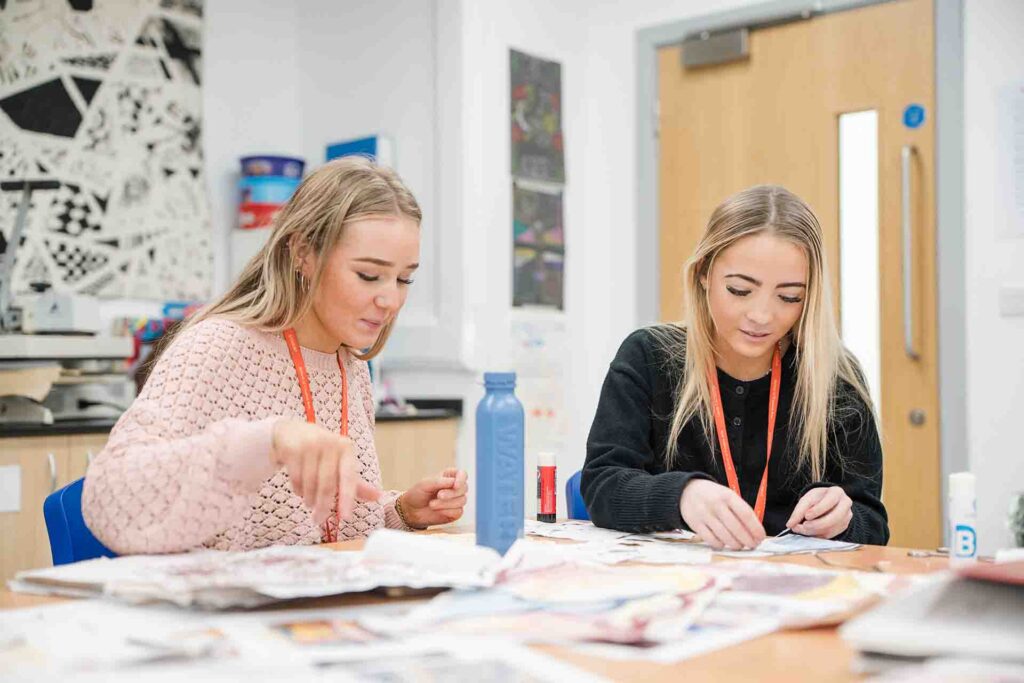 Two female students wearing student lanyards sitting at a desk together in a classroom, crafting with paper snippets and glue sticks, with a blue water bottle on the table between them