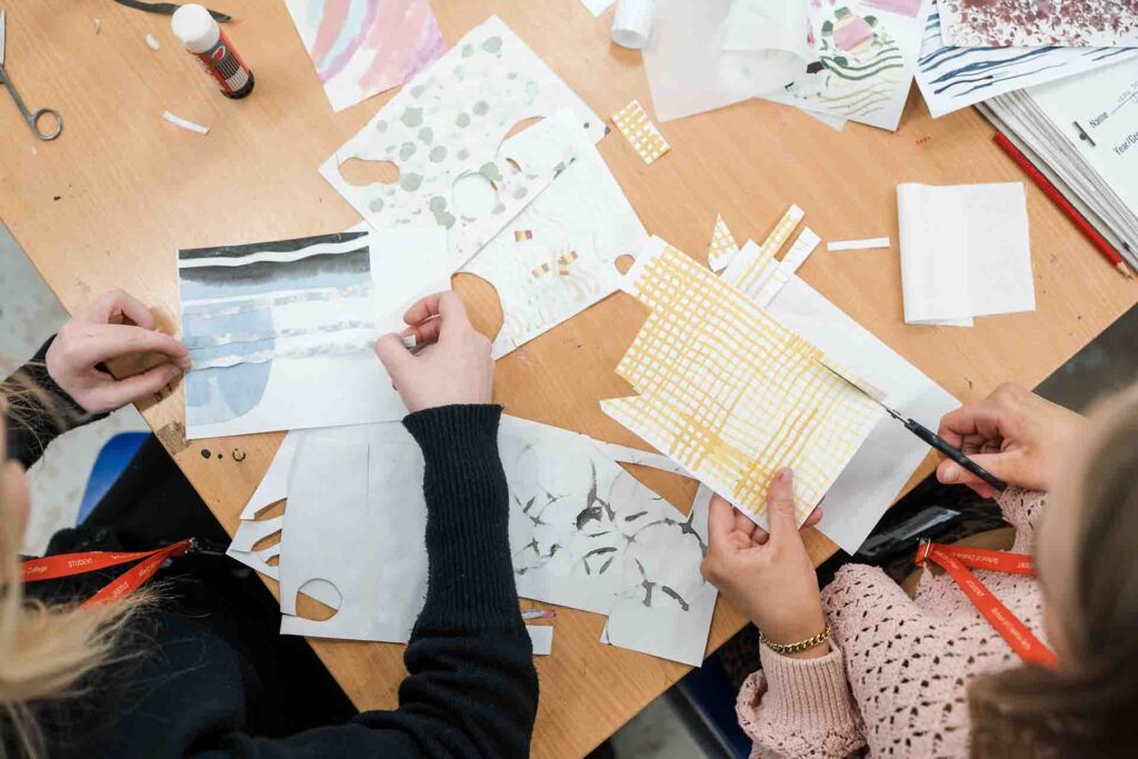 Top-down shot showing the hands of two students collaborating on a paper collage project using scissors, glue, and various patterned, cut-out paper designs spread across a wooden table