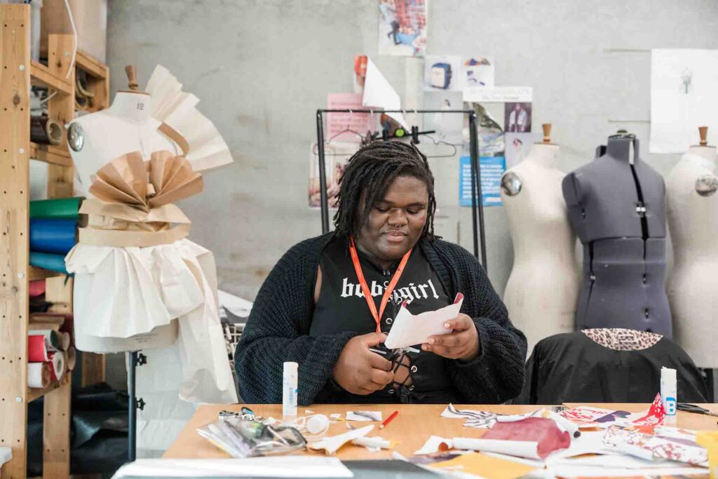 A student with dreadlocks, wearing a black cardigan, black top, and an orange lanyard, carefully cutting paper with scissors at a creative workspace surrounded by dress mannequins, paper designs, and art supplies