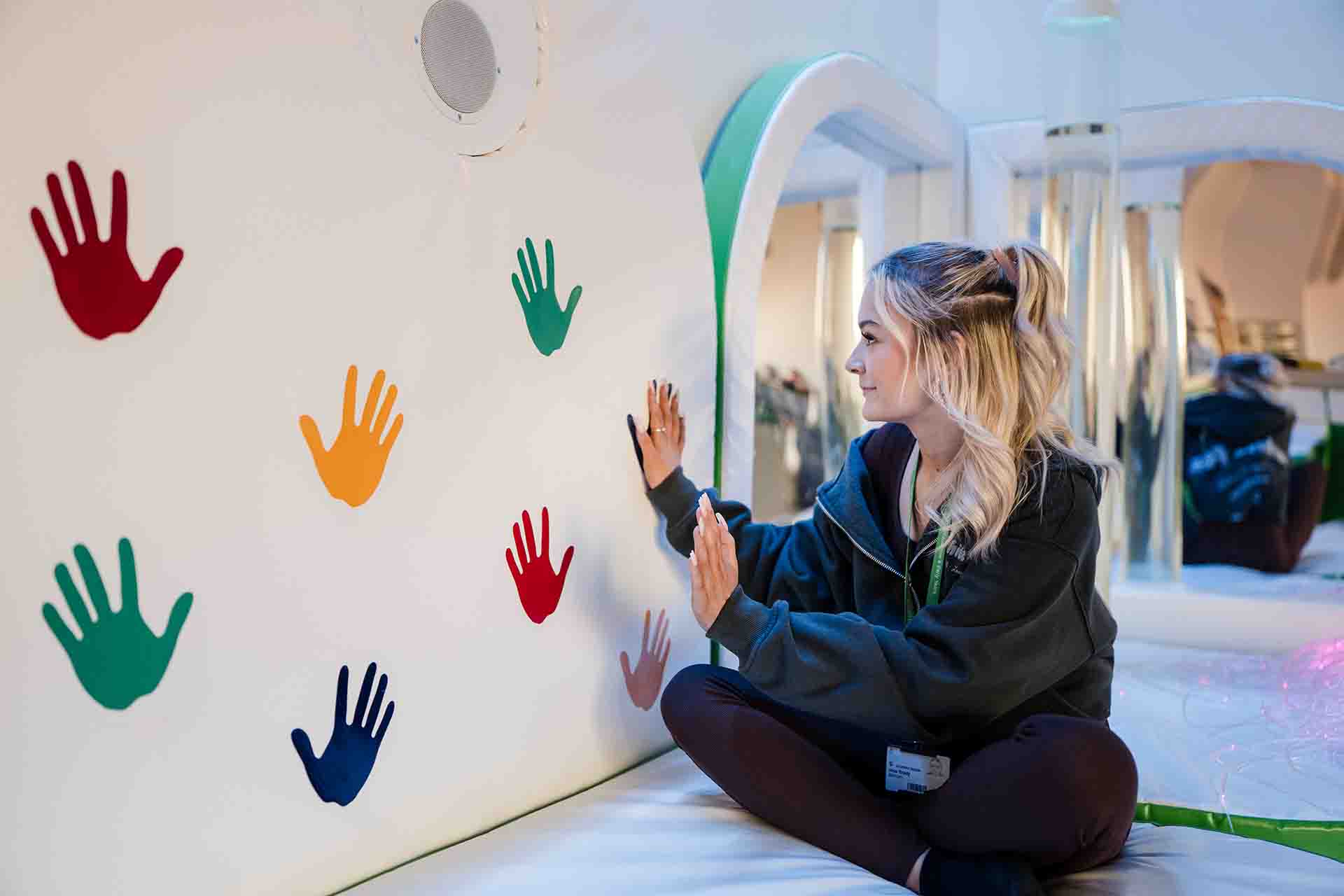 A blonde female student sitting cross legged on a floor, touching a white padded wall decorated with colourful handprint cutouts in a sensory room
