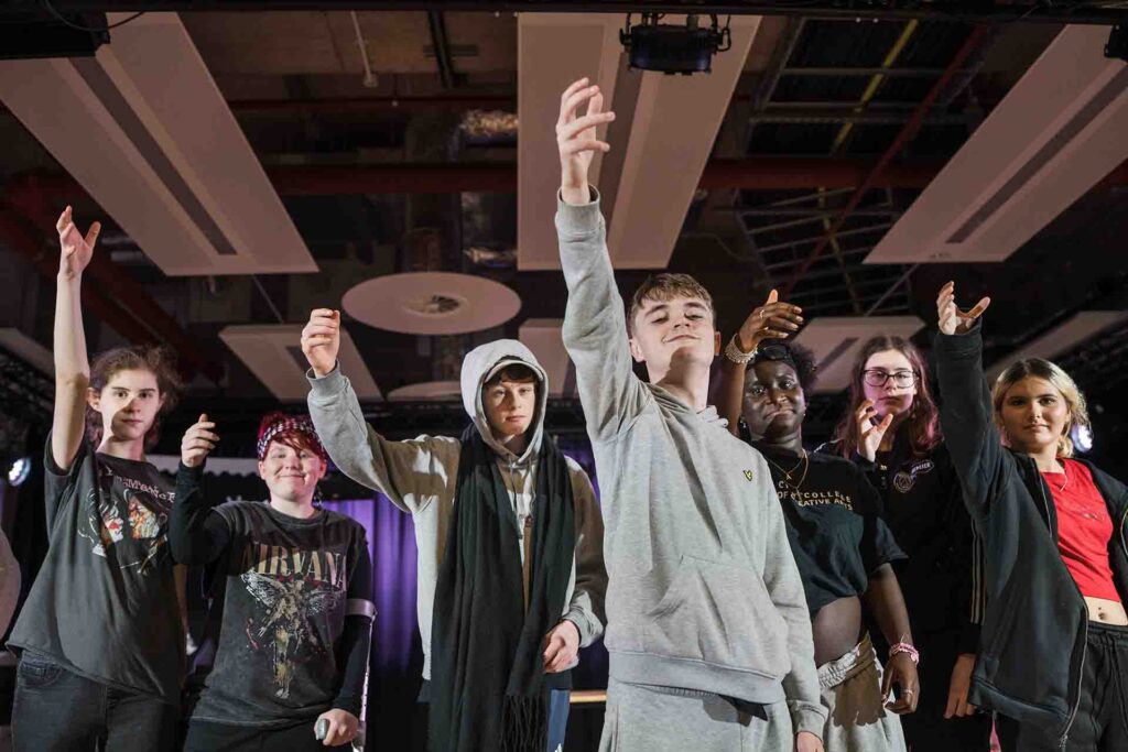 A group of Creative Arts Performing Arts students standing on stage, smiling and raising their right hands while performing