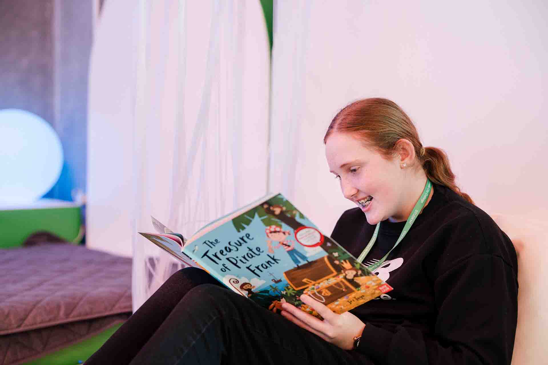 A female student with blonde hair tied back in a ponytail wearing a black sweater, smiling as she reads an open picture book titled 