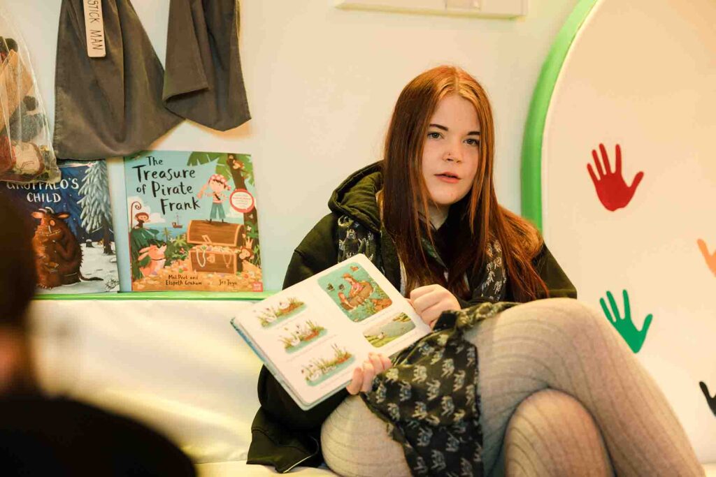 A female student with long reddish brown hair sitting down and holding open a children's picture book in front of a shelf displayed with storybooks