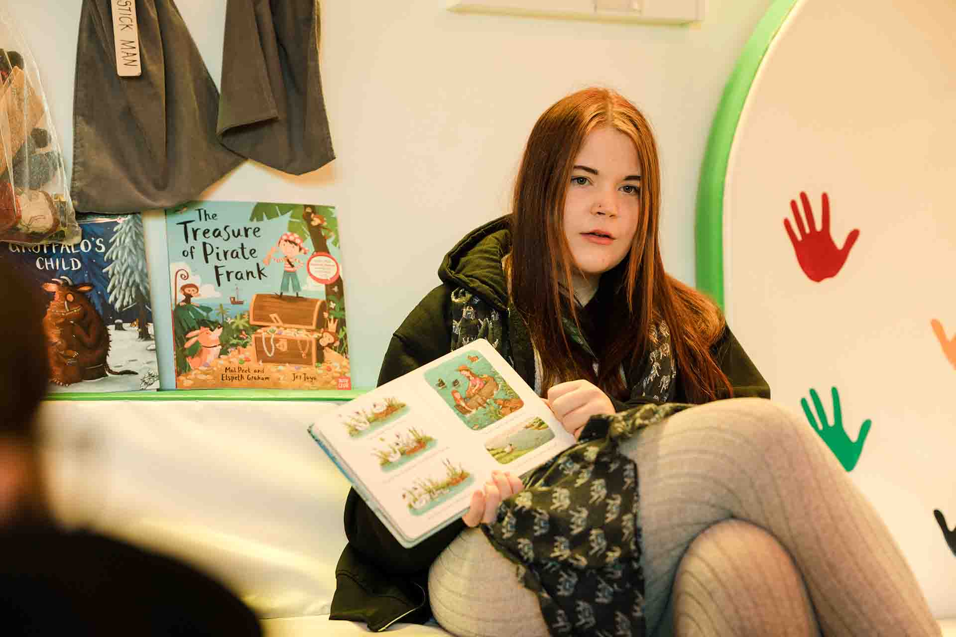 A female student with long reddish brown hair sitting down and holding open a children's picture book in front of a shelf displayed with storybooks