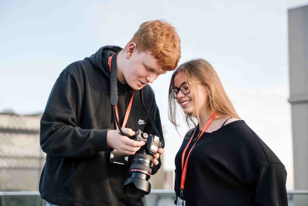 A male student showing photos on his camera screen to a smiling female student wearing glasses outdoors on a rooftop