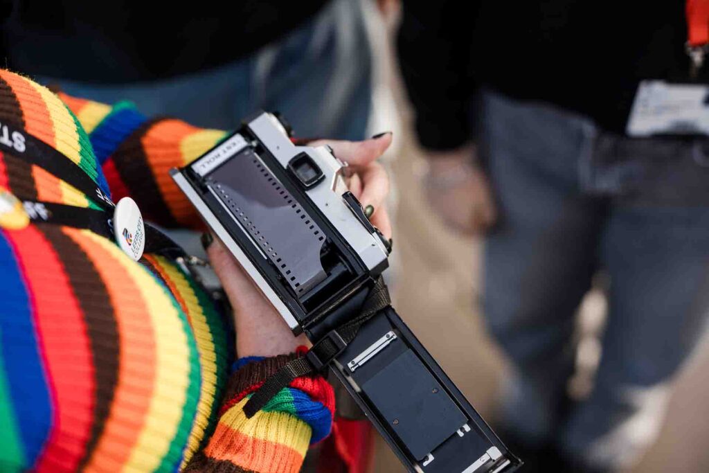 Close-up top view of someone wearing a colourful striped sweater holding an open 35mm film camera, showing the internal film compartment