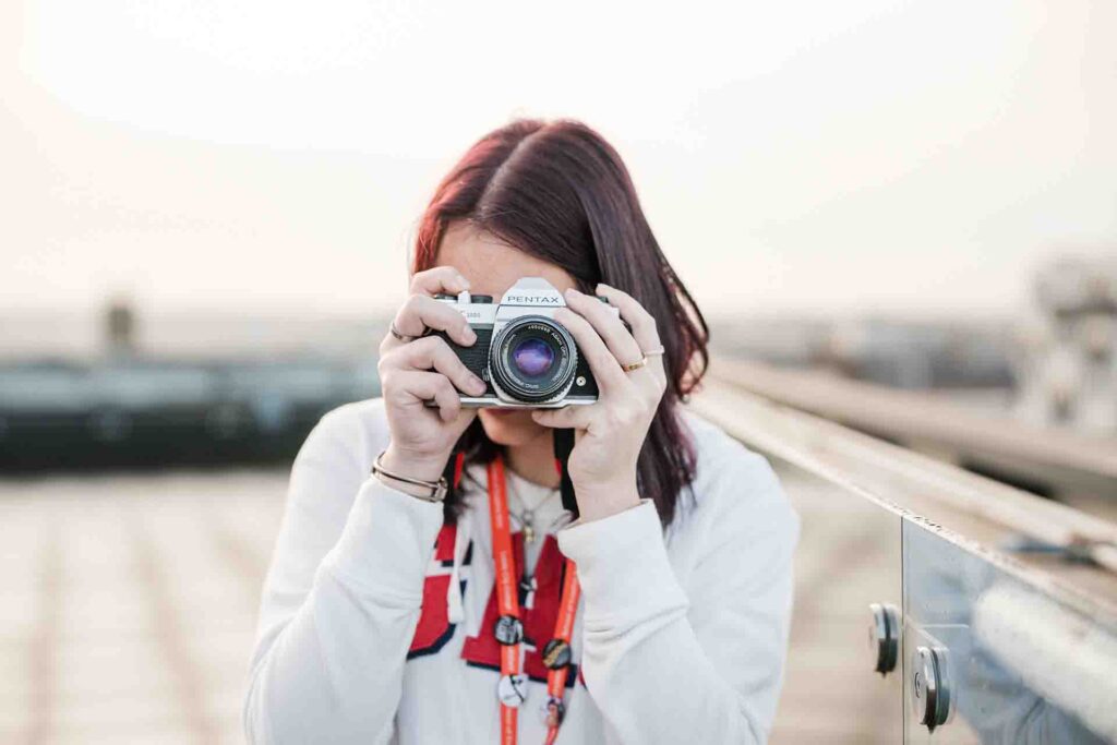 A student wearing a white hoodie holding a vintage silver Pentax film camera up to her face, pointing the lens directly toward the viewer outdoors