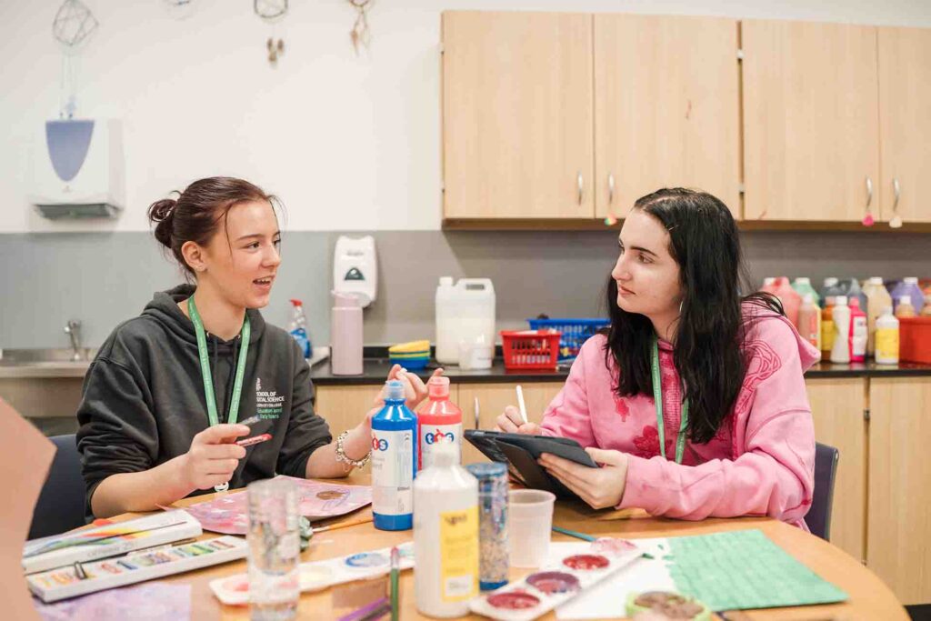 Two female students sitting at a round wooden table in an art room chatting. One wears a dark grey hoodie, and the other wears a pink hoodie while holding a digital tablet. Paint bottles and watercolour trays sit between them