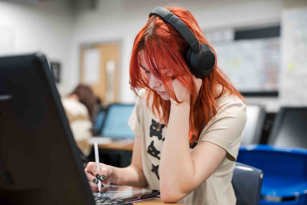 A student with red hair is seated in the classroom, wearing headphones and sketching on her iPad.