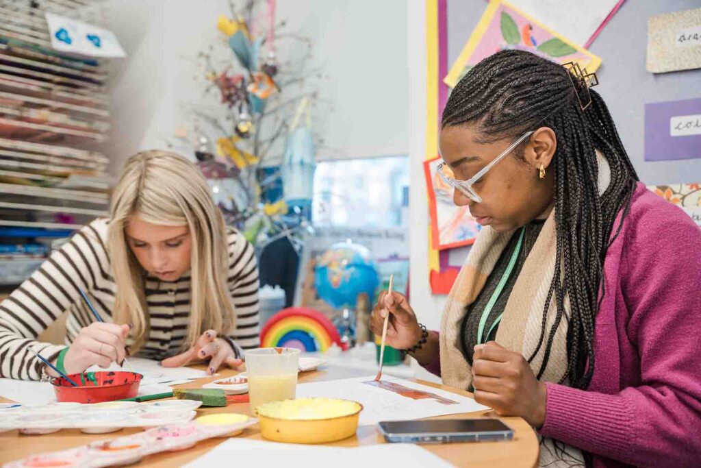 Two students sitting side by side painting on paper at a table. On the right, a young woman with braided hair and glasses paints with a fine brush, while a blonde student in a striped sweater paints on the left