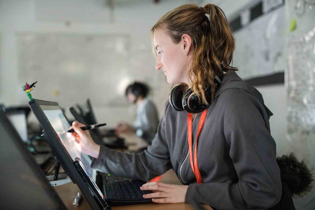 Student in a grey hoodie with a red lanyard and headphones around her neck sketching on a digital screen