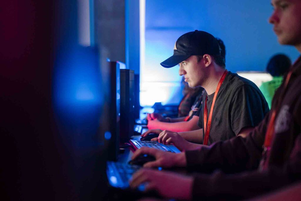 Students sitting in a dimly lit classroom, with one student in a baseball cap concentrating on the screen