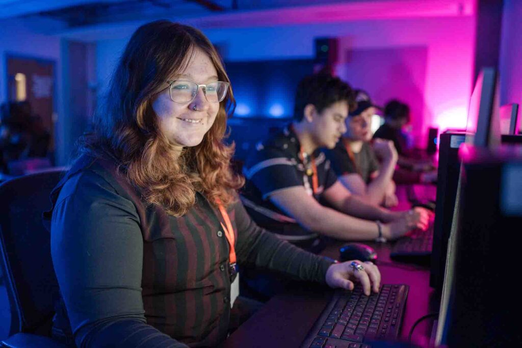 In a dimly lit classroom full of students, a female student is sitting with a big smile on her face as she concentrates on her computer screen