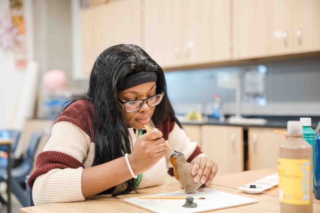 A female student with glasses, a black headband, and a striped sweater carefully applying paint with a fine brush to a small clay sculpture at an art studio worktable