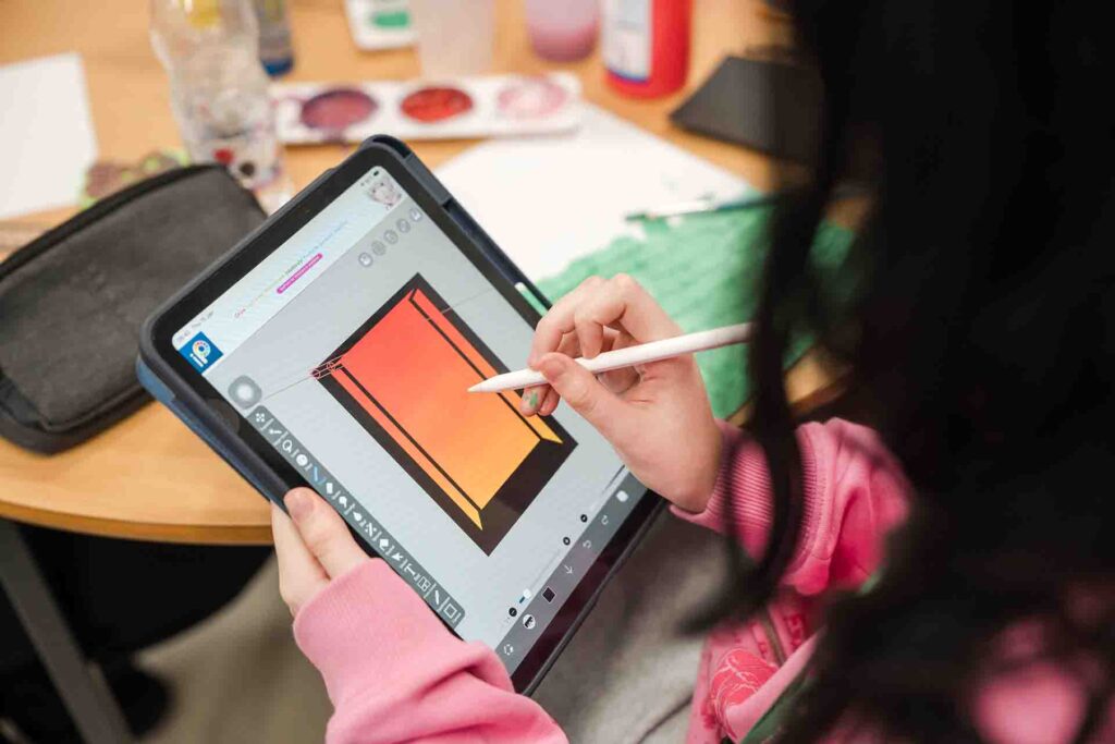 Over the shoulder shot of a student wearing a pink hoodie using a stylus to draw a 3D rectangular prism outline on a digital tablet at a table filled with art supplies
