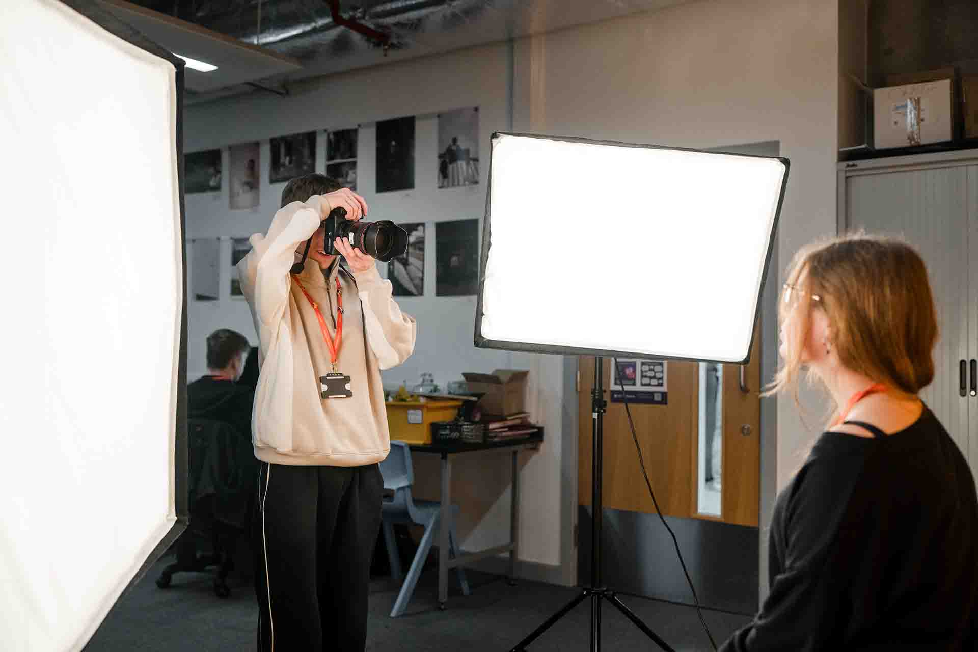 A young photographer holding a DSLR camera to their eye while taking a portrait of a female student in a studio setup with bright soft box lights