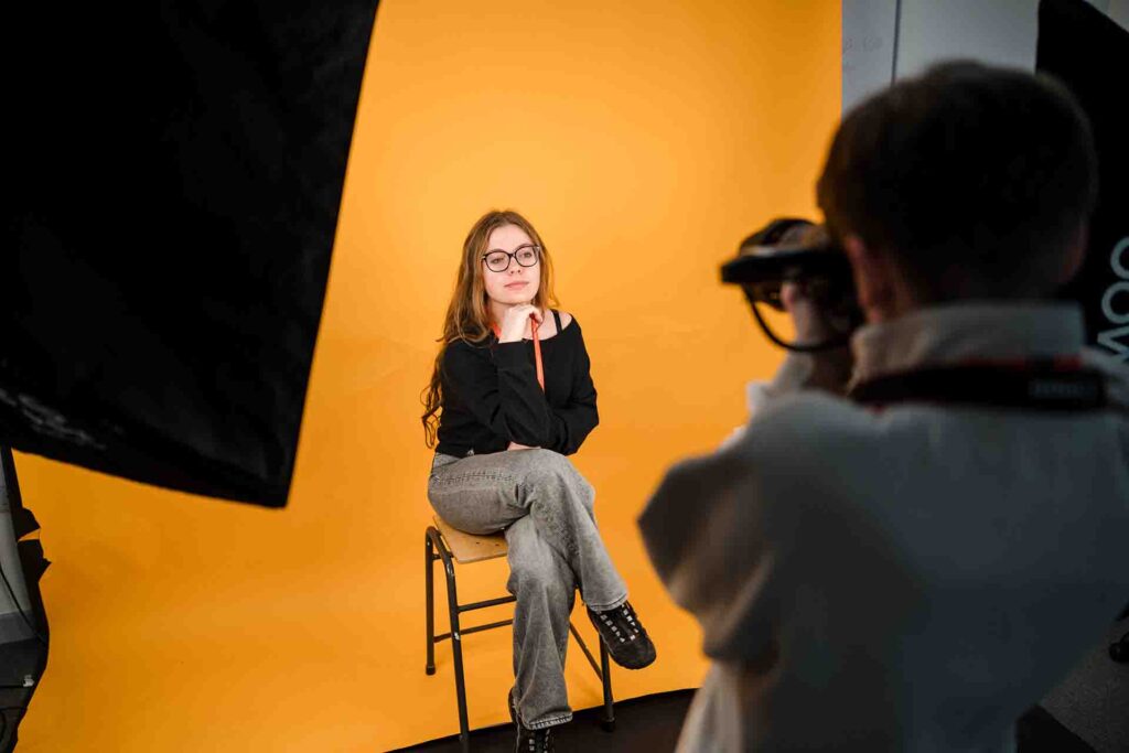 A young woman with glasses posing on a stool against a bright yellow studio backdrop while a photographer in the foreground takes her picture