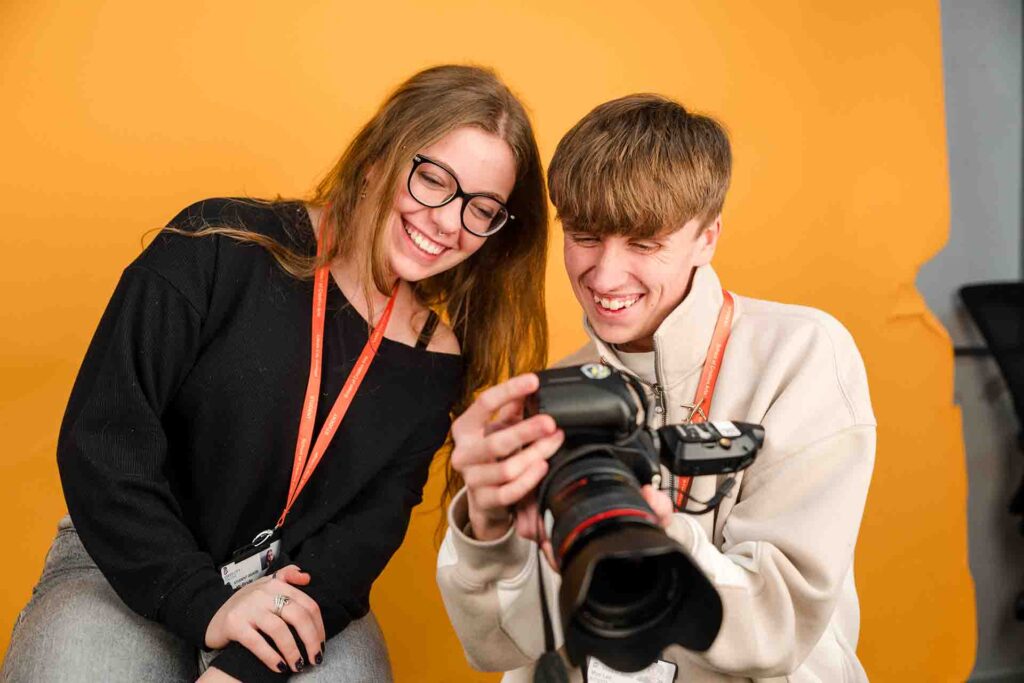 Two smiling students leaning together to review photos on the digital screen of a DSLR camera in front of a yellow studio backdrop