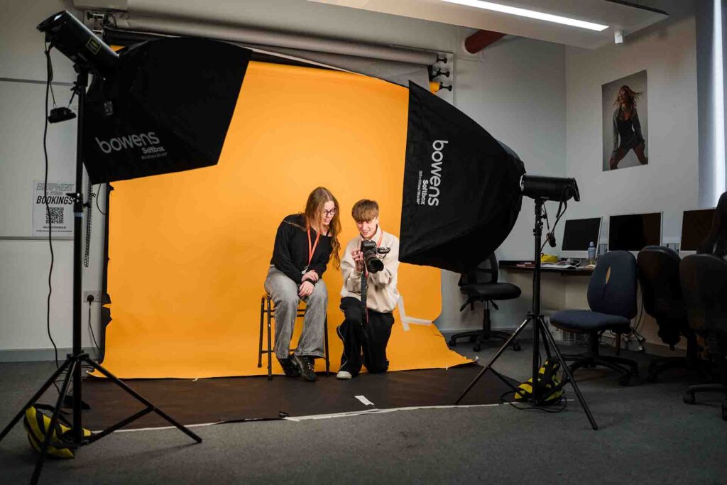 Wide shot of two students reviewing photos on a camera inside a photography studio equipped with large Bowens soft box studio lights and a yellow backdrop