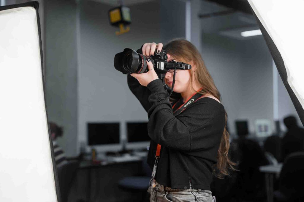 Medium close-up of a female student holding a large Canon camera up to her eye, taking a shot in a studio environment
