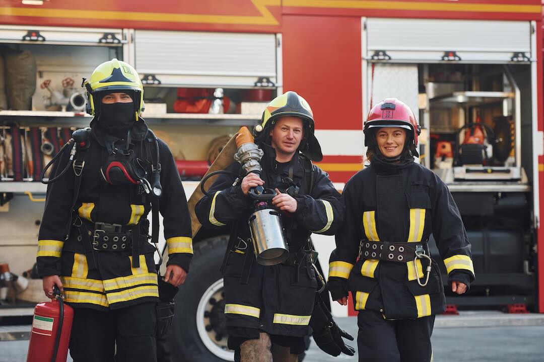Three firemen walking away from the fire truck carrying a hose and a fire extinguisher