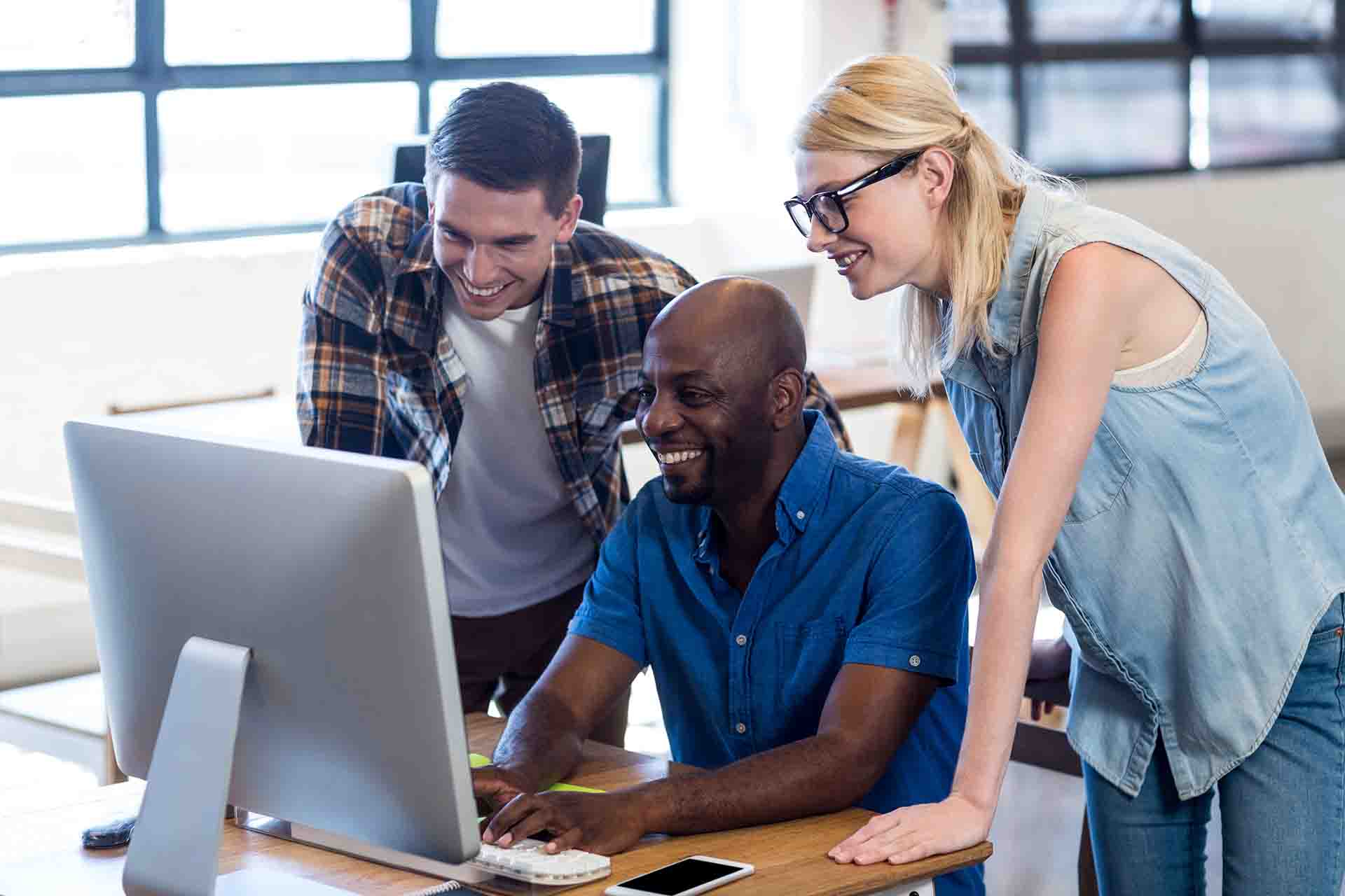 Three diverse colleagues smiling together in an office while leaning over to look at a desktop computer monitor on a wooden desk
