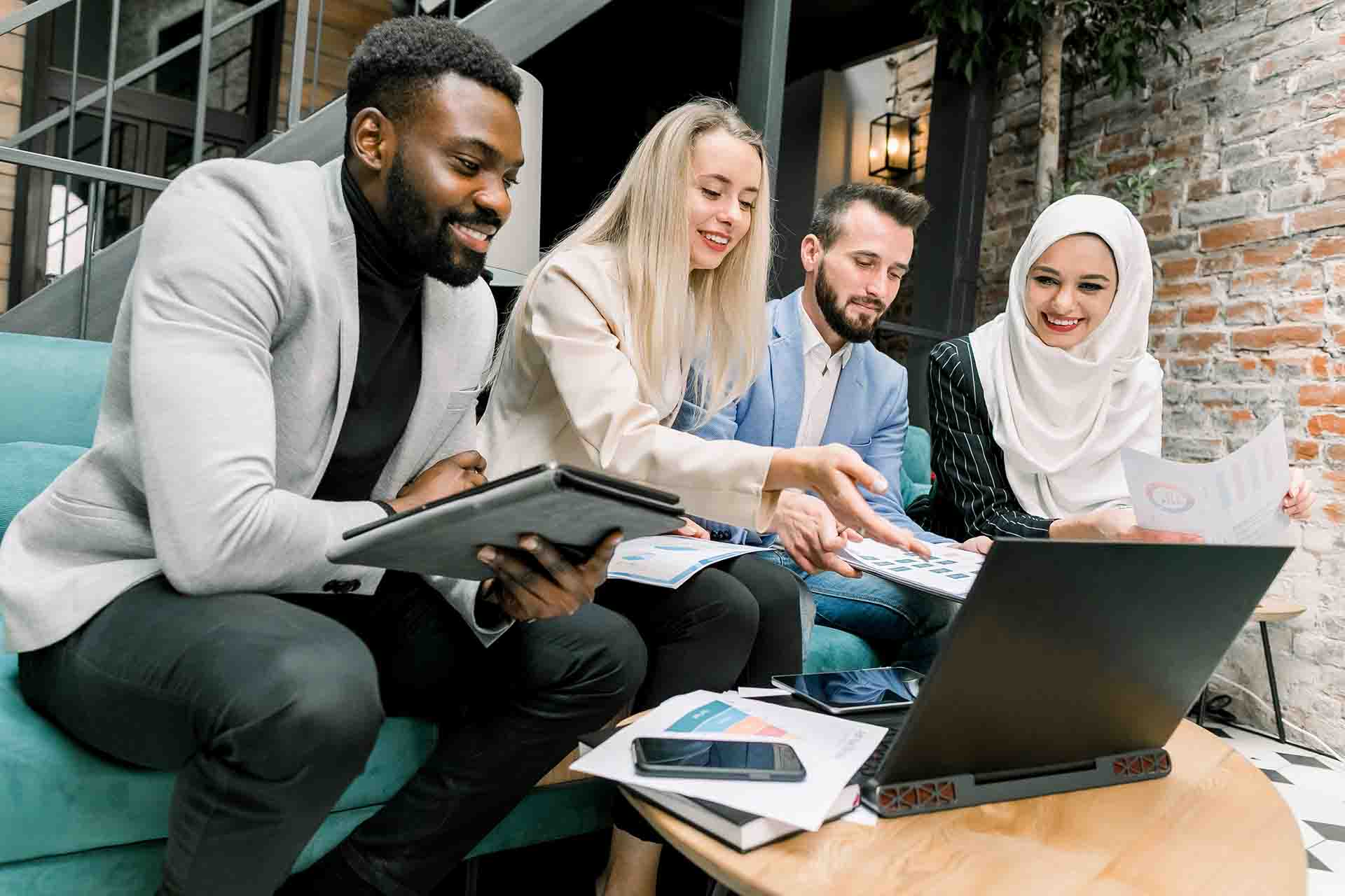 Four diverse business professionals sitting on a couch around a coffee table covered with papers, pointing at a laptop screen during a group discussion