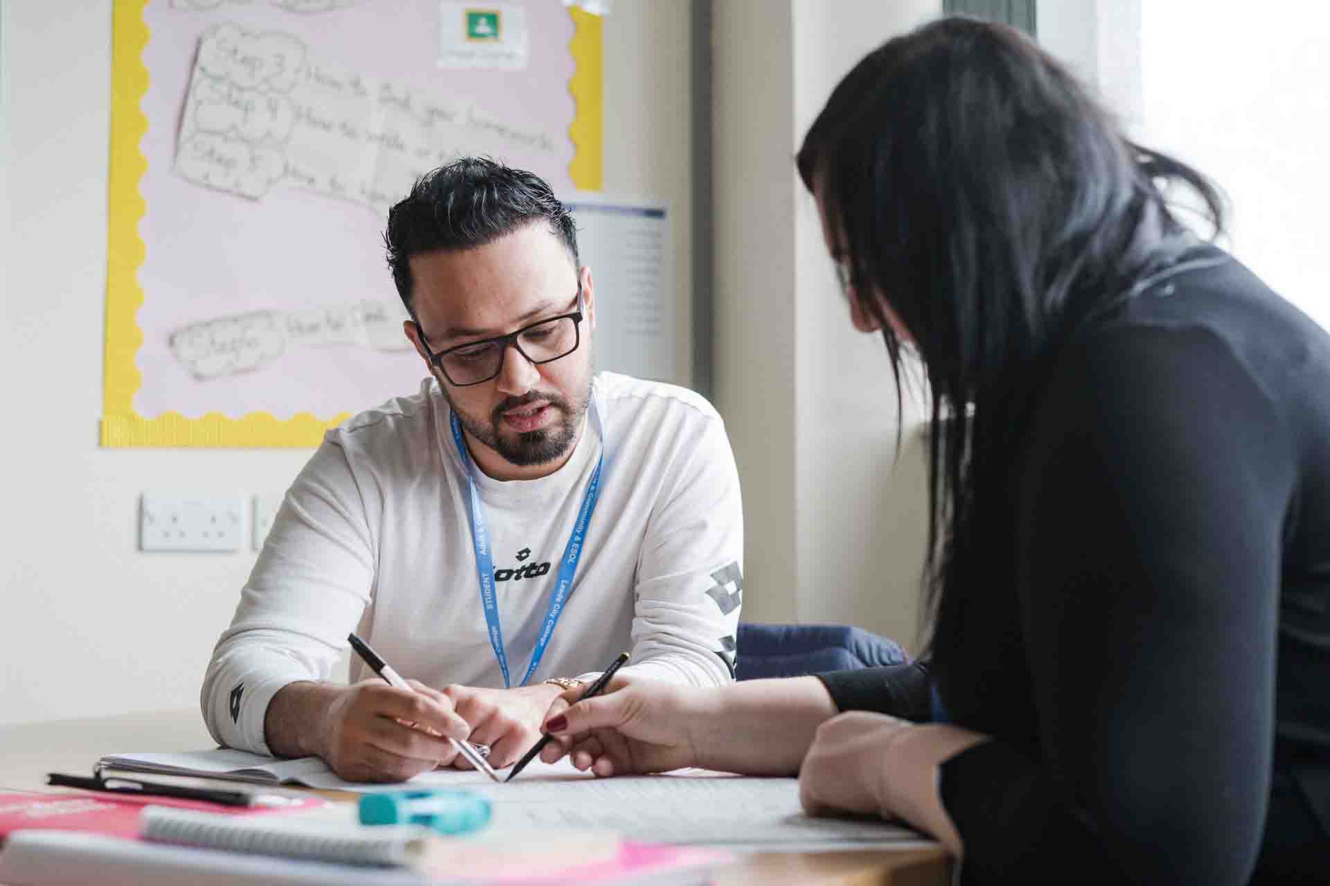 A man with glasses wearing a Leeds City College lanyard works closely with an instructor at a desk, both pointing at a shared worksheet