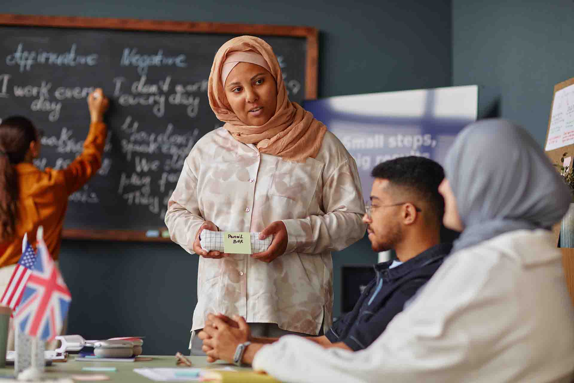 A teacher wearing a peach hijab speaks to a classroom of adult students while holding a small pencil box labeled with a sticky note. In the background, another student writes on a chalkboard displaying grammar notes