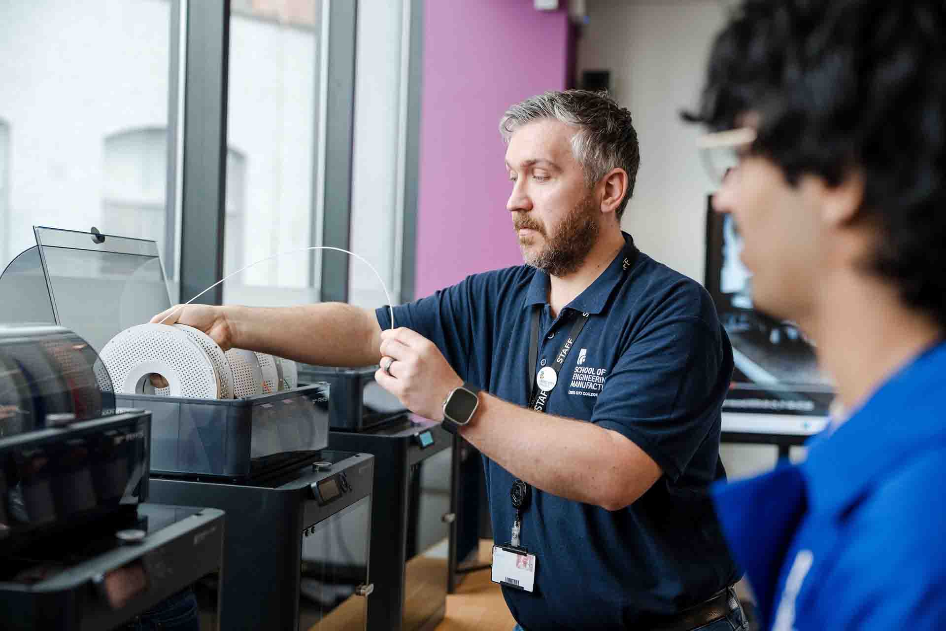 A staff member wearing a dark blue polo shirt demonstrates how to load a filament spool into a 3D printer for a student standing nearby