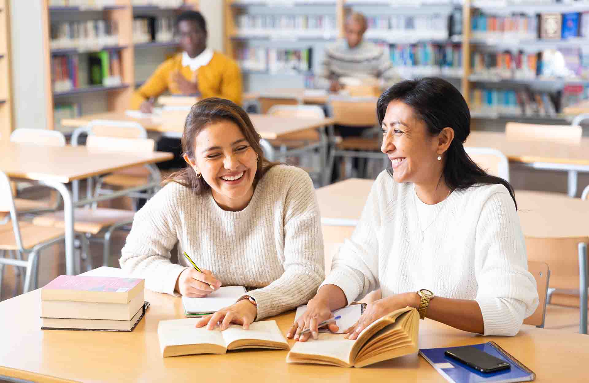 Two women sit together smiling and laughing at a desk in library, studying open textbooks with notebooks and pens