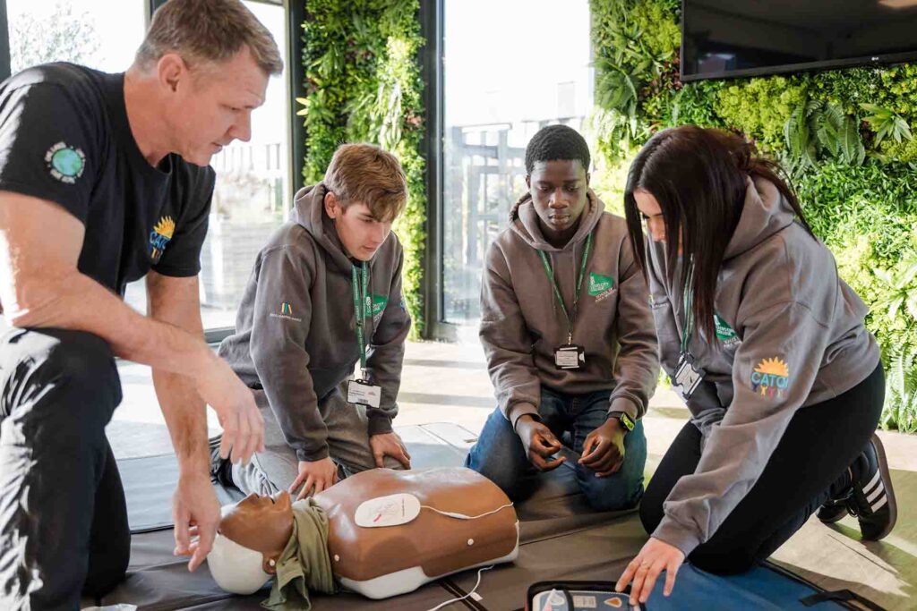 An instructor and three Leeds City College Public Services students kneeling on the floor around a CPR training torso, practicing first aid and defibrillator setup.