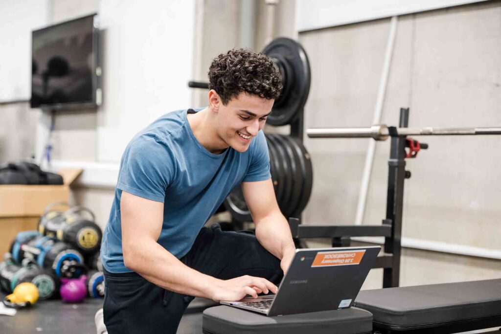 A smiling young man kneeling next to a weight bench in a gym setting, typing on a laptop rested on the bench pad