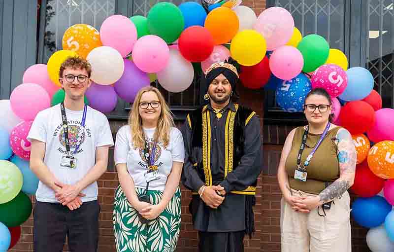 Four smiling individuals standing beneath a large, colourful balloon arch during a college of sanctuary event