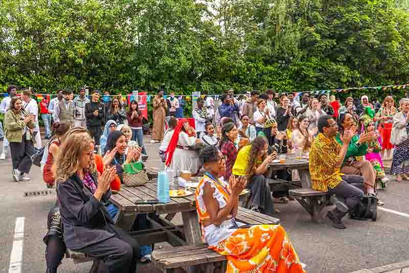 An enthusiastic crowd seated outdoors at picnic tables clapping at a college of sanctuary event