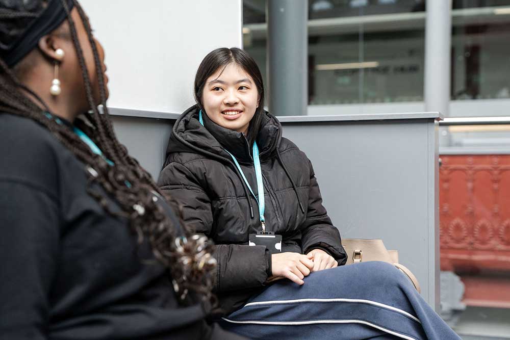 Two students wearing college lanyards sitting on a bench and chatting indoors.