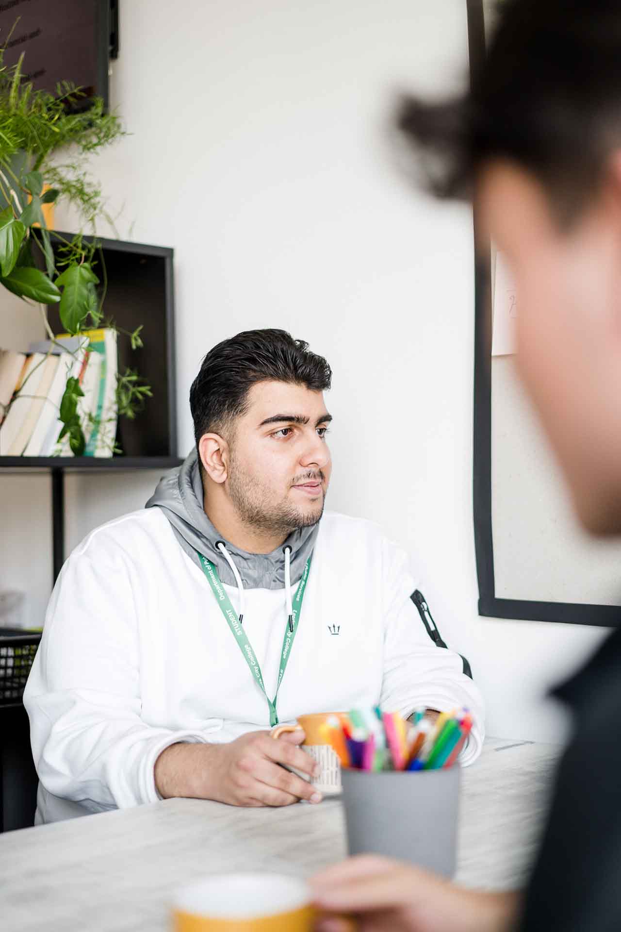 A student in a white hoodie sitting at a desk with a warm drink, talking to a classmate