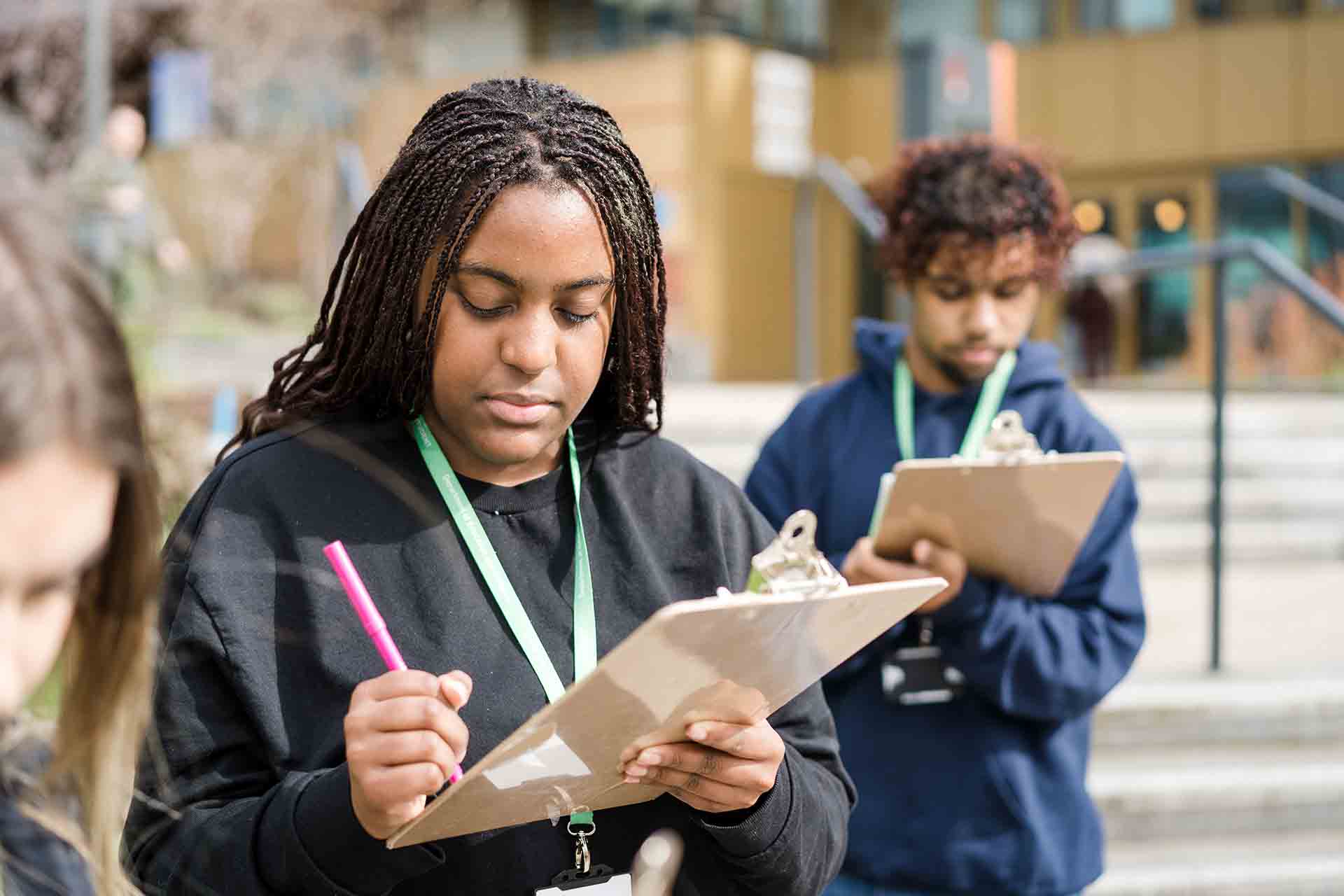 A student with long dreadlocks taking notes on a clipboard outdoors with classmates