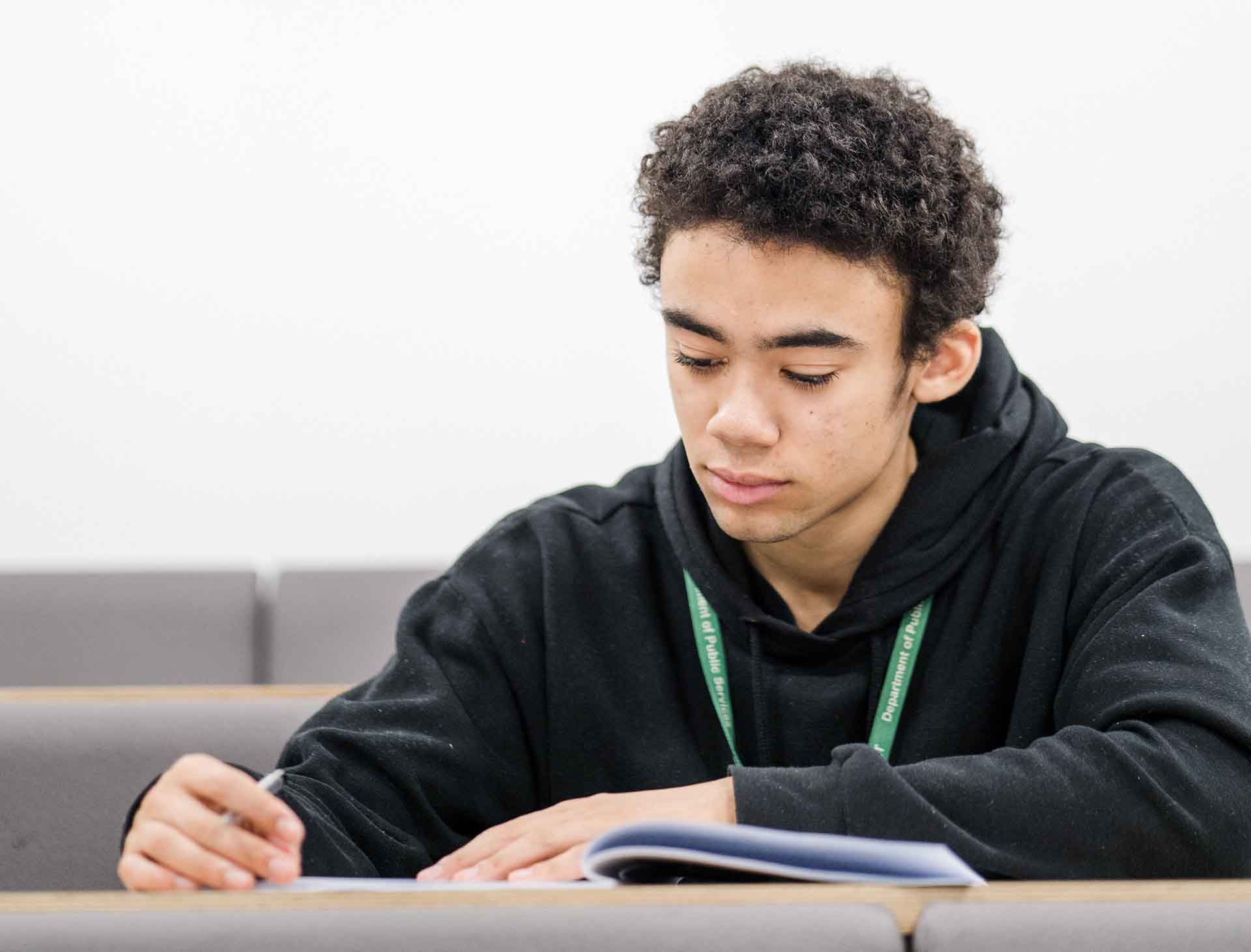A student sitting at a desk wearing a black hoodie and green lanyard, writing in a notebook