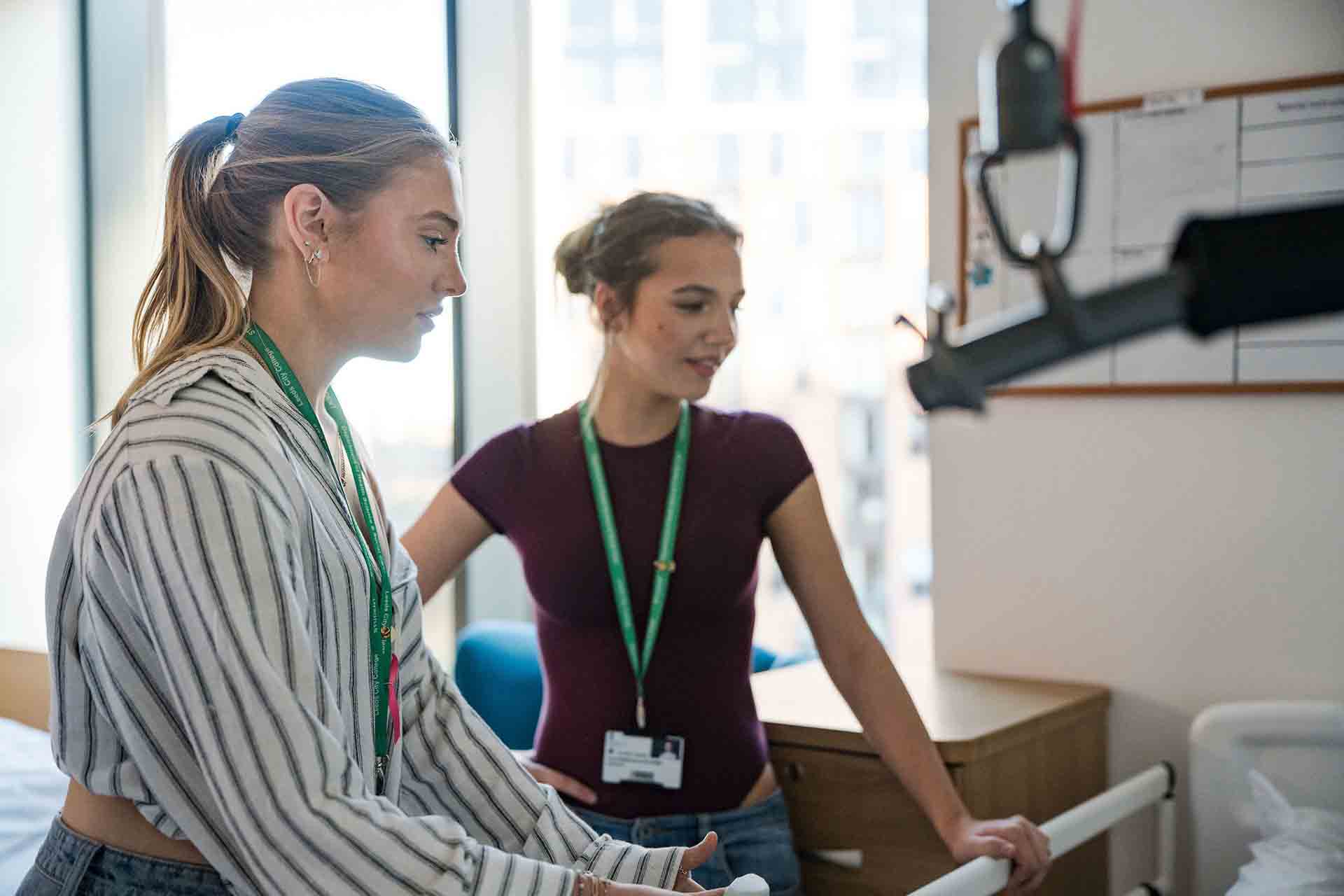 Two Leeds City College students wearing green lanyards working with medical training equipment near a hospital bed setup.
