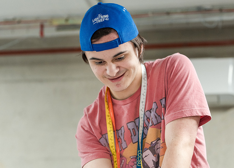 Student with cap on backwards and measuring tapes around his neck looking down smiling