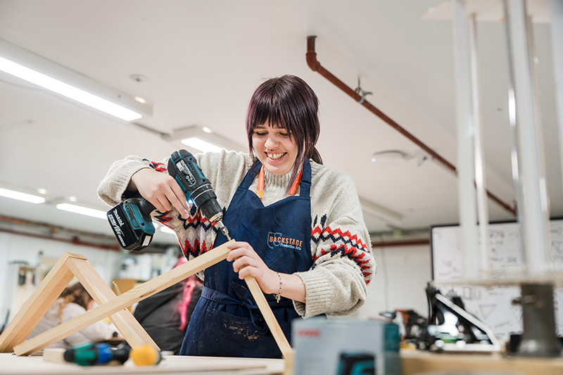 A person is stood smiling using a screwdriver to fit together two pieces of wood for a stage set