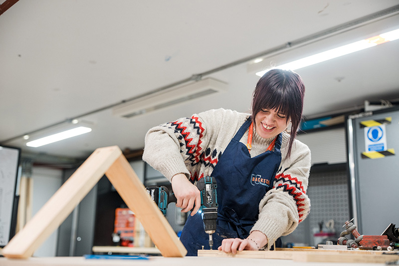 A person is stood smiling using a screwdriver to fit together pieces of wood for a stage set