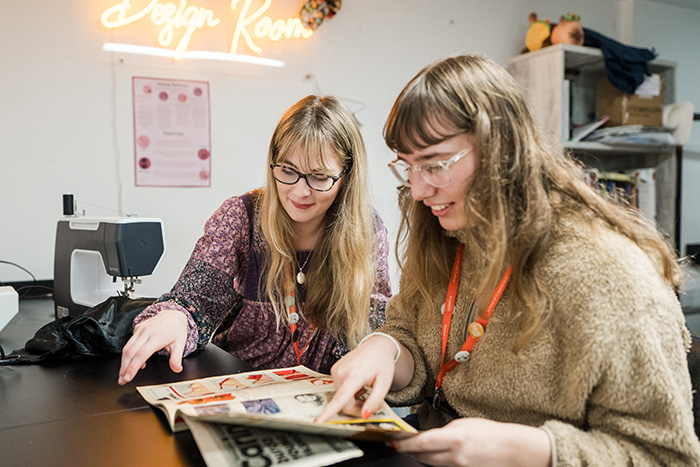 Two people sat next to a sewing machine in the design room looking at fabric patterns in a book