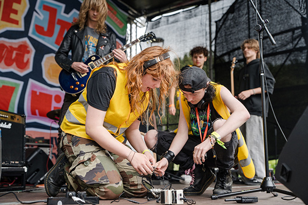 Two students with high vis jackets working on sound equipment on stage next to musicians with guitars