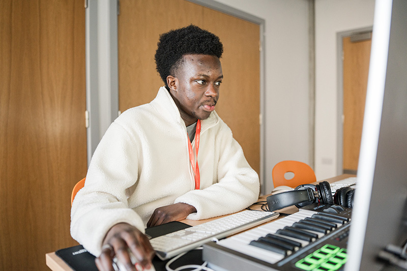 Person sat working on music technology with headphones and keyboard next to a desktop screen and mouse